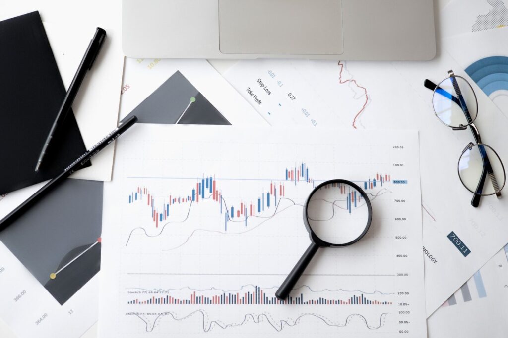 Magnifying glass resting on printed financial charts beside pens, reading glasses and a laptop on a white desk.