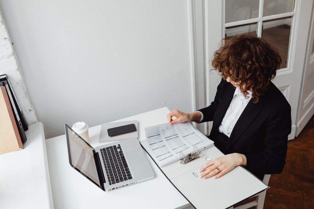 Woman in a black blazer reviewing printed documents in a binder beside a laptop at a white desk.