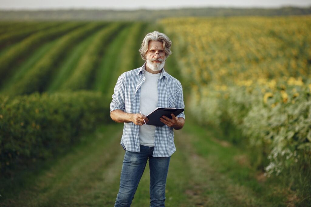 Grey-haired farmer in a checked shirt holding a folder and pen on a track between green crop rows and a sunflower field.