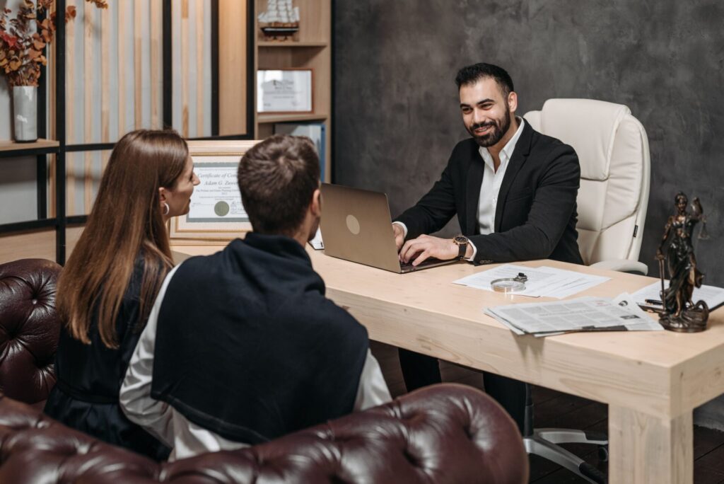 Lawyer at a desk with a laptop and paperwork talking with a couple seated in leather armchairs during a client consultation.