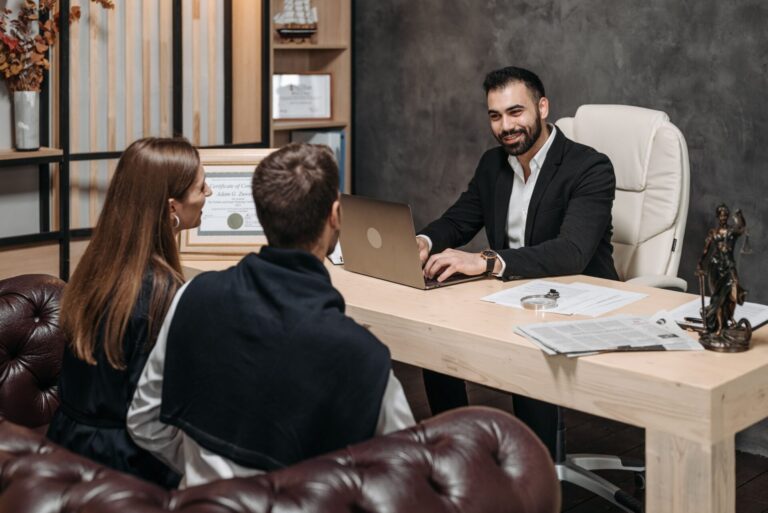 Lawyer at a desk with a laptop and paperwork talking with a couple seated in leather armchairs during a client consultation.