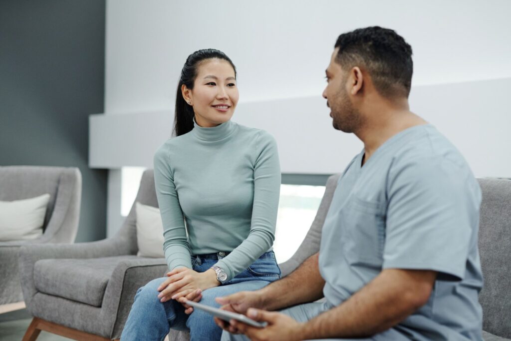 A clinician in scrubs holding a tablet talks with a smiling patient on a grey couch in a modern clinic.