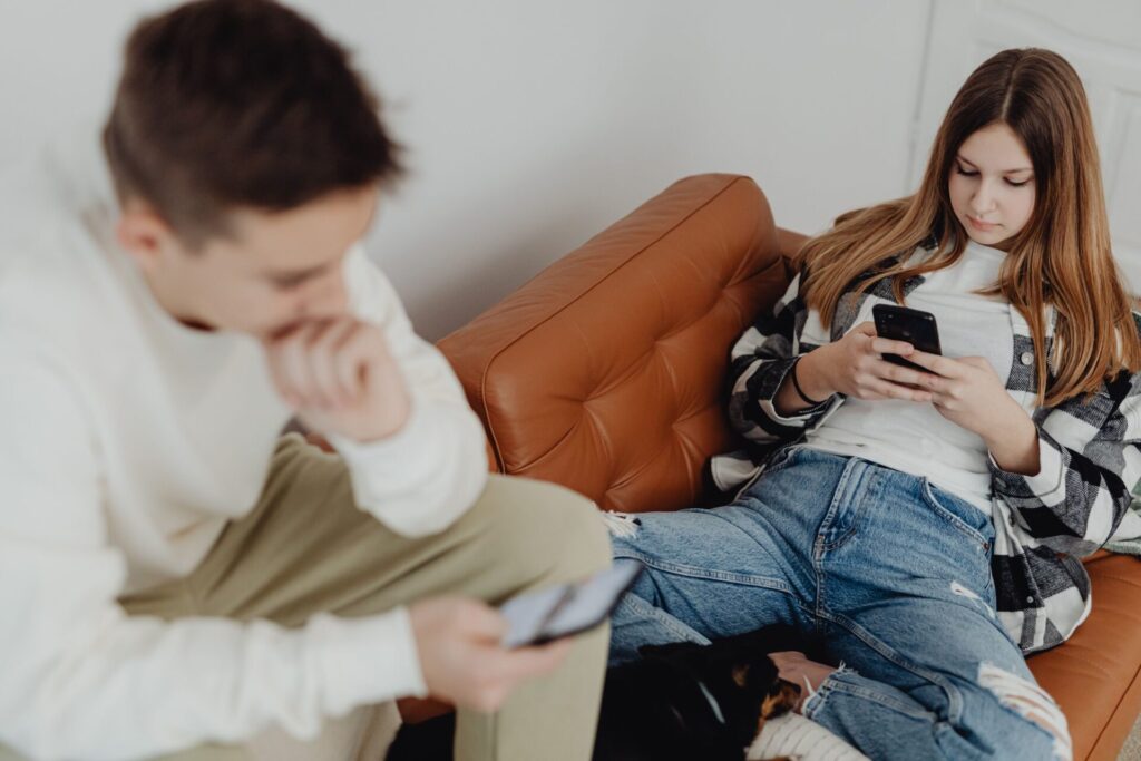 Two teenagers sitting on a tan leather couch at home, each looking at a smartphone.