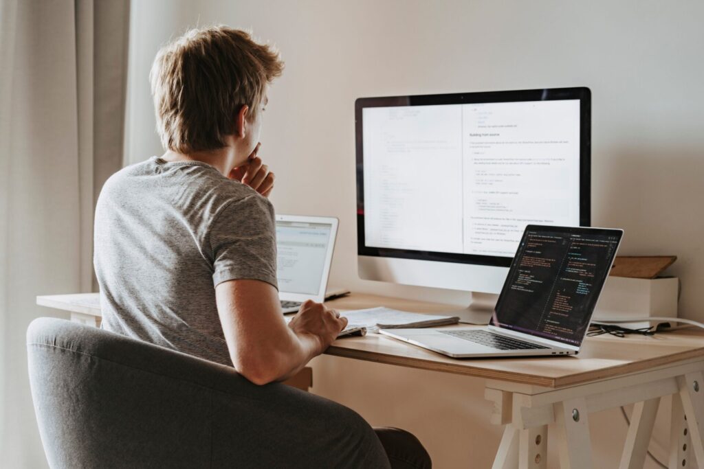 Software developer at a wooden desk reviewing code on a laptop and documentation on a large desktop monitor, with a second laptop beside him.