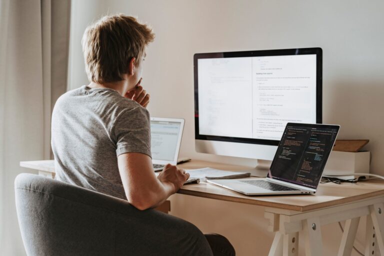 Software developer at a wooden desk reviewing code on a laptop and documentation on a large desktop monitor, with a second laptop beside him.